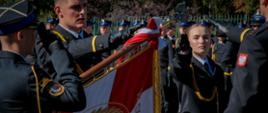 Zbliżenie na moment ślubowania – funkcjonariuszka PSP w czarnym galowym mundurze z naszywką PSP i złotym sznurem dotyka sztandaru szkoły z godłem Polski, wykonując gest przysięgi.