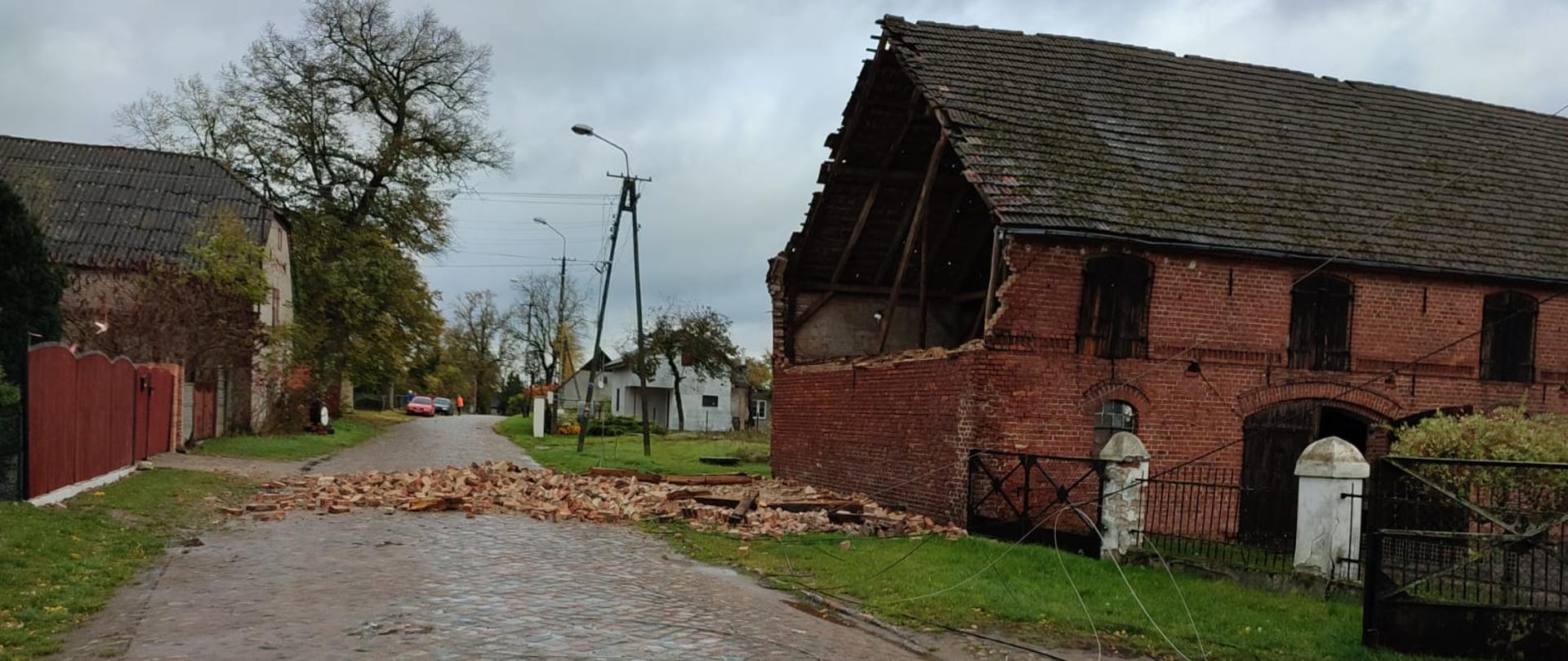 Zdjęcie przedstawia budynek gospodarczy z czerwonej cegły w którym zawaliła się ściana szczytowa. W tle zdjęcia znajdują się inne zabudowania.
