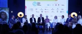 Uczestnicy panelu siedzą na krzesłach w półokręgu na scenie i dyskutują. Za ich plecami wyświetlona jest plansza z nazwą wydarzenia, organizatorami i partnerami. 