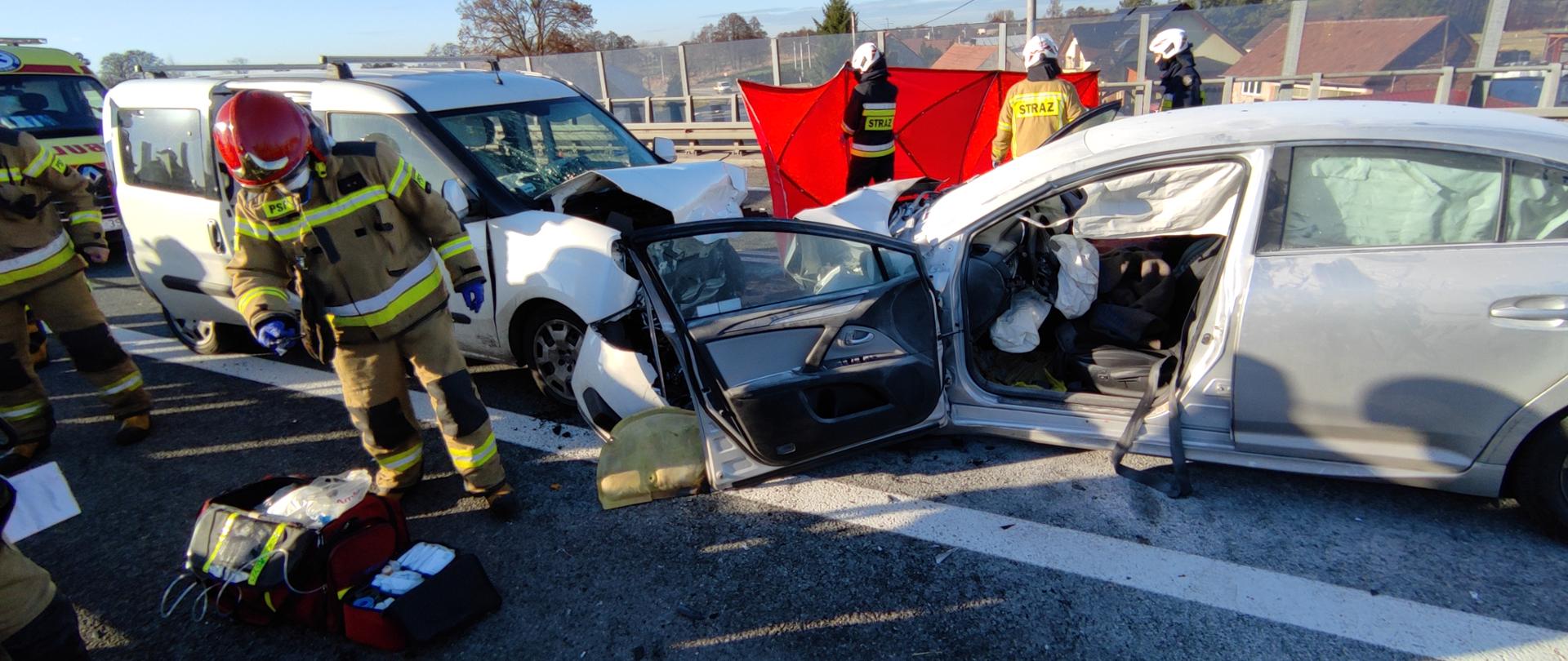 Zderzenie dwóch samochodów osobowych, strażacy w ubraniach specjalnych i hełmach.
