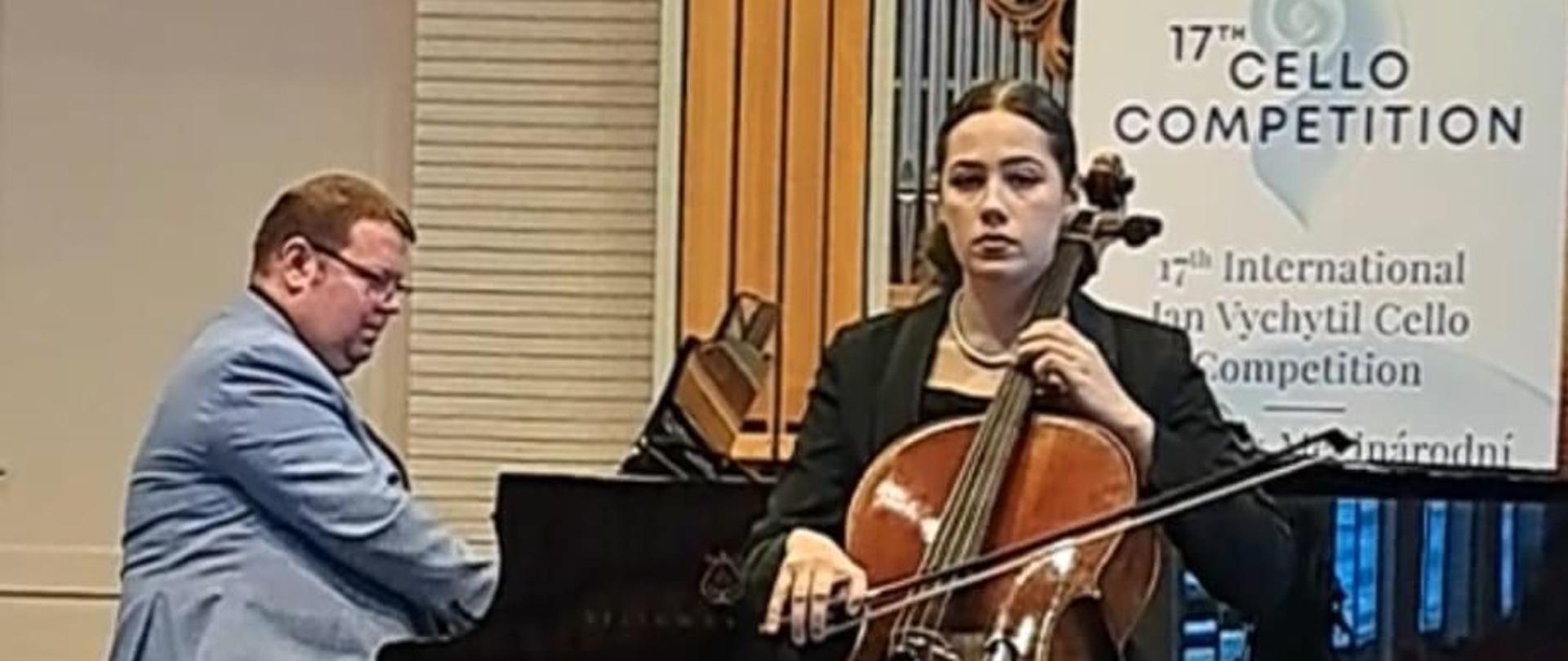 Nastolatka gra na wiolonczeli, za Nią na fortepianie gra mężczyzna, z tyłu widać baner reklamowy konkursu.