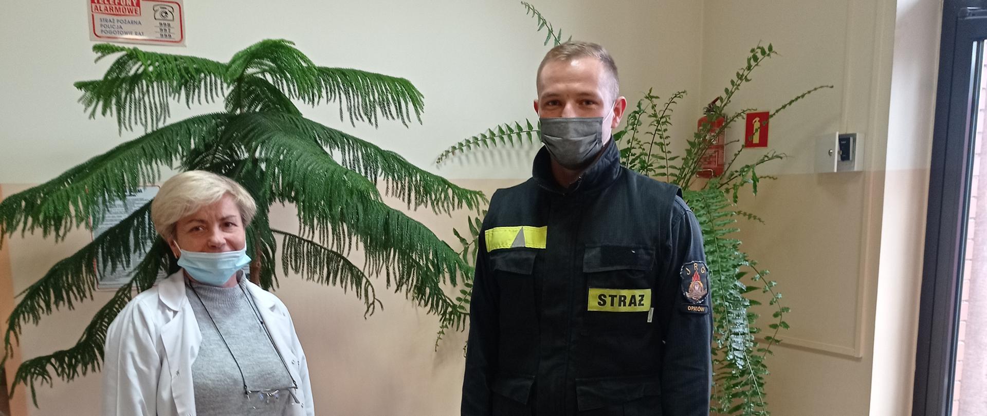  Zdjęcie przedstawia strażaka w ubraniu koszarowym przekazującego środki ochrony indywidualnej przedstawicielce punktu szczepień, oboje w maseczkach chirurgicznych, na podłodze stojące pudła ze SOI