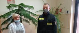  Zdjęcie przedstawia strażaka w ubraniu koszarowym przekazującego środki ochrony indywidualnej przedstawicielce punktu szczepień, oboje w maseczkach chirurgicznych, na podłodze stojące pudła ze SOI