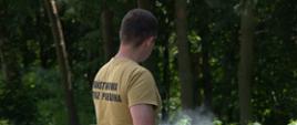 Funkcjonariusz ubrany w koszulkę z napisem „Państwowa Straż Pożarna” demonstruje działanie środka gaśniczego w terenie leśnym; w tle widoczne jest stanowisko do kontrolowanego ognia.