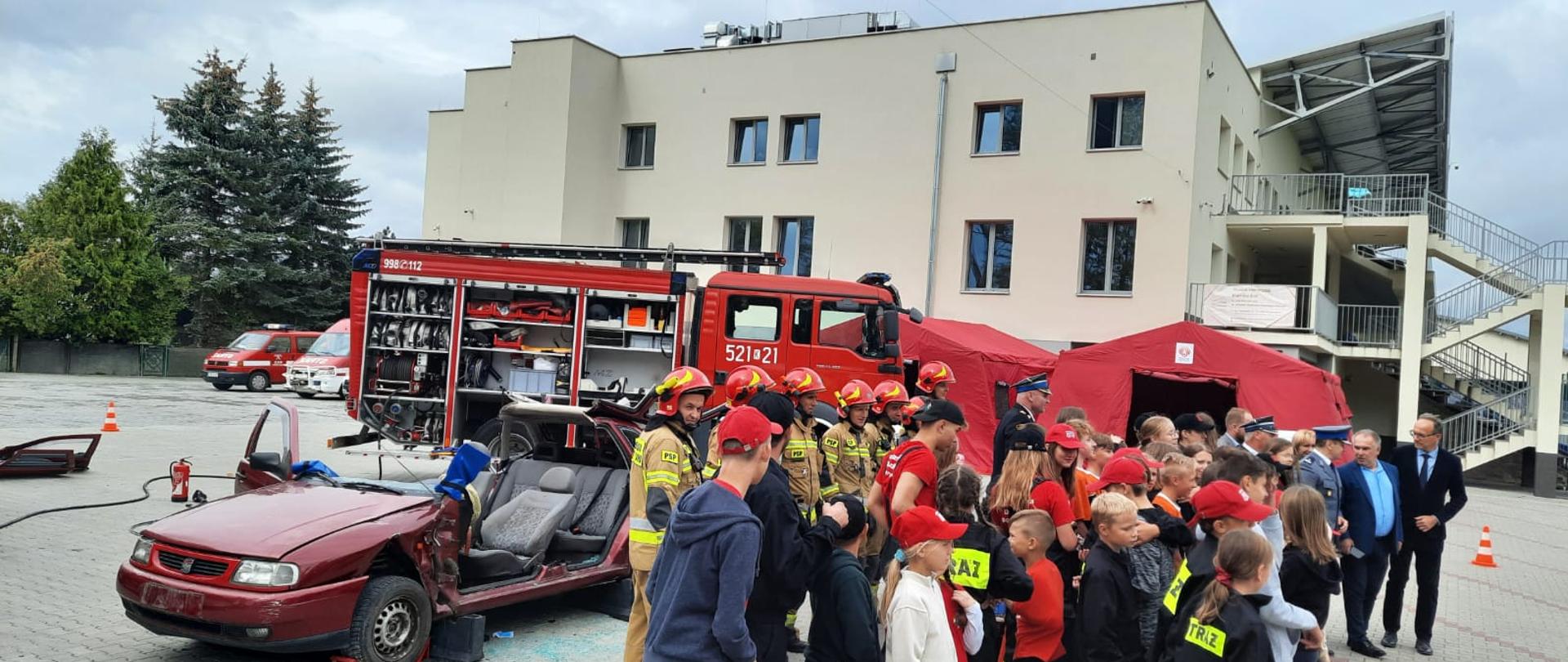 Grupa dzieci przygląda się strażakom jak tną pojazd