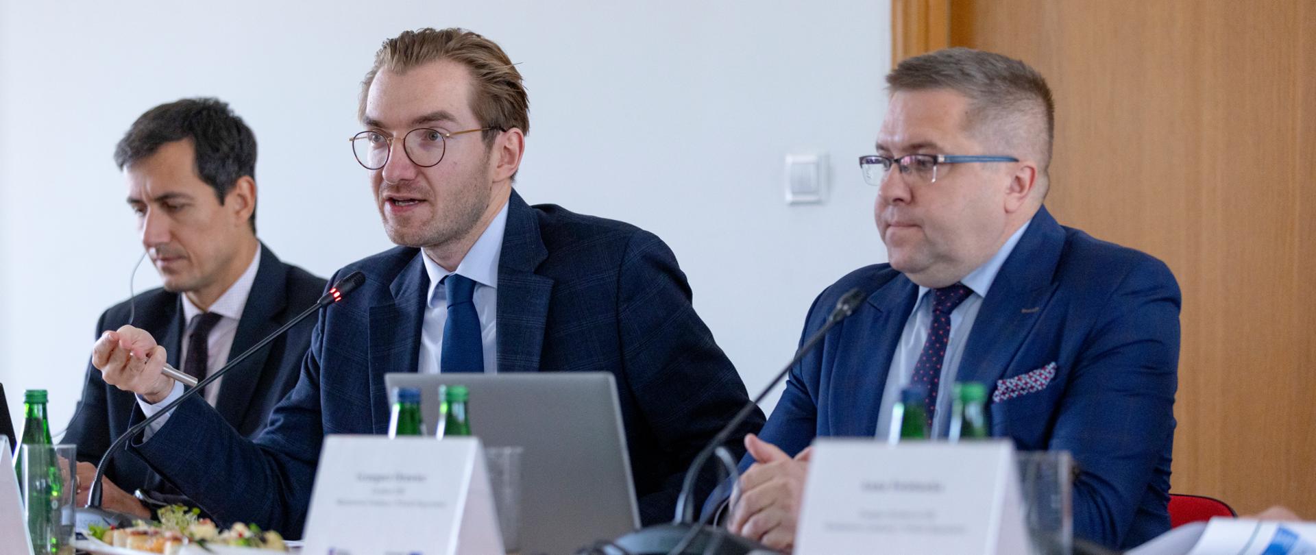 Trzech mężczyzn siedzi przy stole zastawionym przez mikrofony, laptopy i butelki. W środku wiceminister Jan Szyszko.