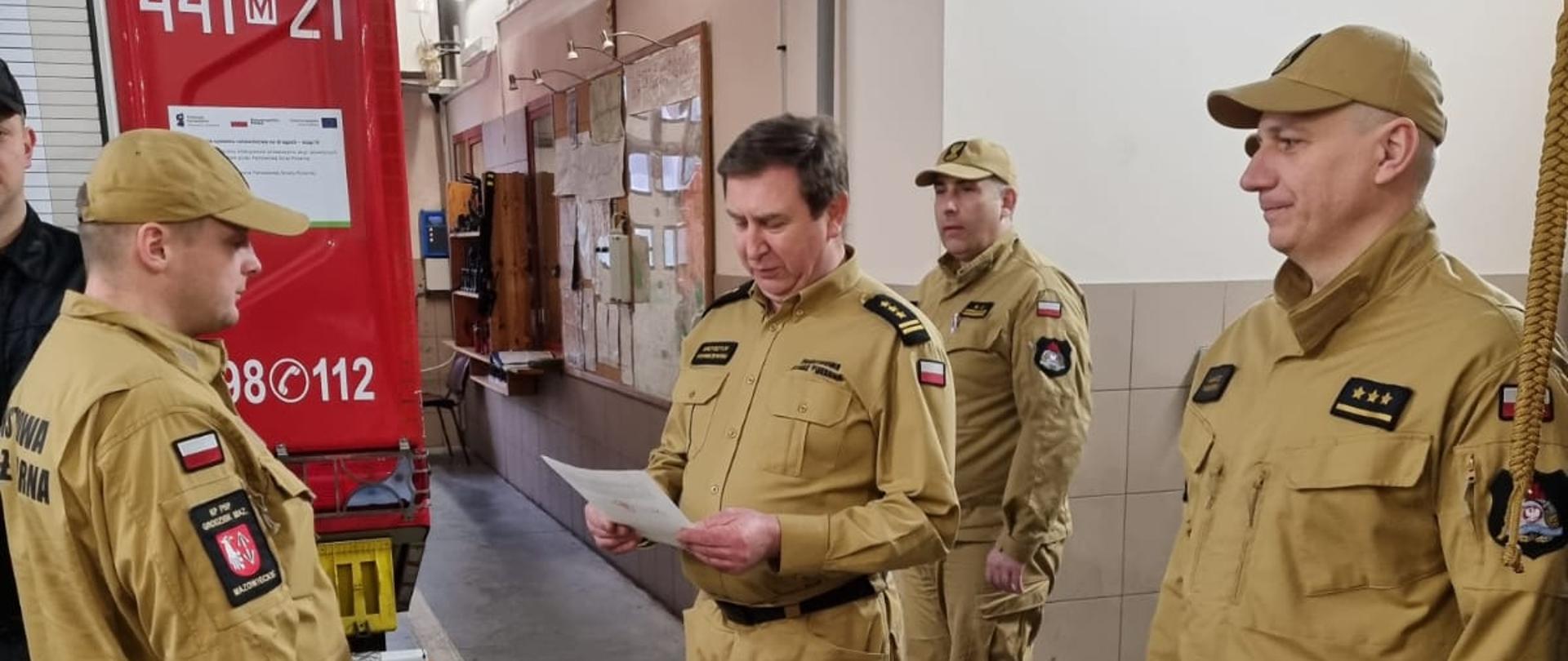 Komendant Powiatowy PSP w Grodzisku Maz. st. bryg. Krzysztof Tryniszewski wręcza mianowanie na stanowisko dowódcy zmiany st. asp. Łukaszowi Mikulskiemu w obecności p.o. Z-cy Komendanta Powiatowego PSP w Grodzisku Ma. bryg. Marka Szymanka, D-cy JRG kpt. Mariusza Wołowskiego, Z-cy D-cy JRGH st. kpt. Bartosza Szulborskiego oraz mian zdającej i przyjmującej w pomieszczeniu garażowym JRG.