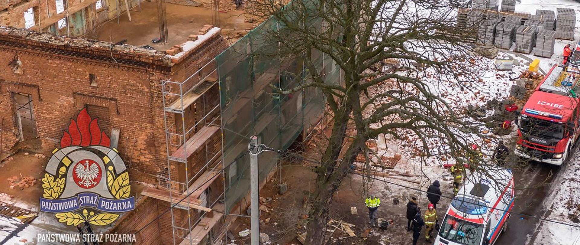 Zniszczony budynek w trakcie remontu, na rusztowaniu przy budynku stoją strażacy, przed budynkiem stoi biała karetka pogotowia oraz czerwony wóz strażacki. W lewym dolnym rogu logo Państwowej Straży Pożarnej