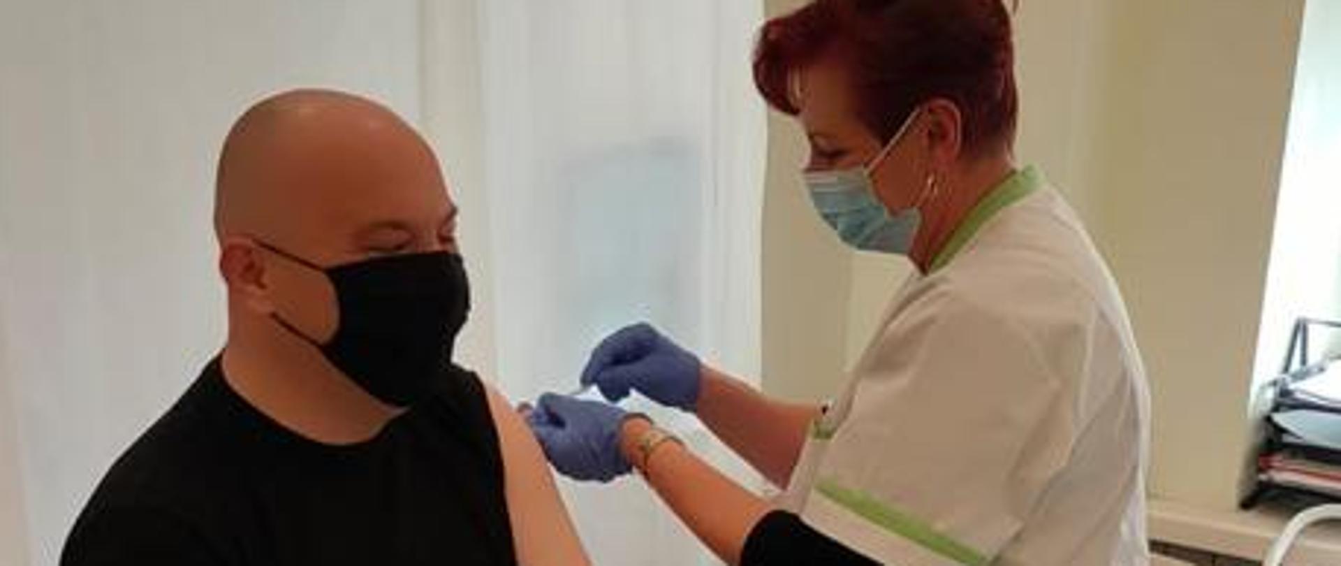 Zdjęcie przedstawia strażak PSP w momencie wykonywania szczepienie przeciwko covid-19. Na tle białego parawanu, na łóżku zabiegowym siedzi strażak ubrany na czarno, któremu pielęgniarka wykonuje szczepienie w lewe ramię. Pielęgniarka ma na sobie biały fartuszek. Oboje mają na twarzach maseczki. 