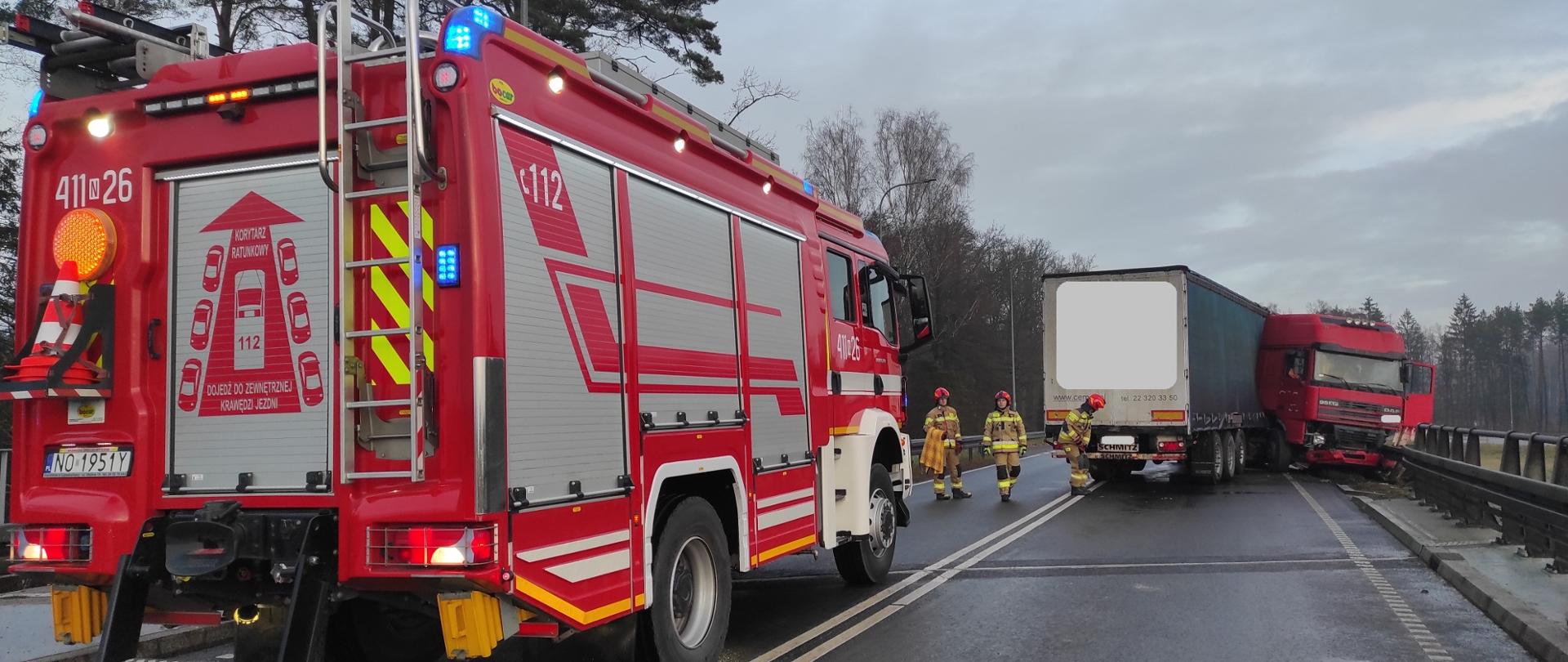 Zdjęcie przedstawia miejsce zdarzenia drogowego z udziałem samochodu ciężarowego, które miało miejsce na drodze dwujezdniowej. Ciężarówka uderzyła w barierę energochłonną, zatrzymując się częściowo poza jezdnią. Na miejscu obecni są strażacy w ubraniach specjalnych, którzy zabezpieczają miejsce zdarzenia oraz prawdopodobnie sprawdzają stan pojazdu.
Na pierwszym planie widoczny jest średni samochód ratowniczo-gaśniczy Państwowej Straży Pożarnej. Na pojeździe oraz w jego otoczeniu aktywne są sygnały świetlne ostrzegawcze (niebieskie i pomarańczowe).
Zdjęcie ukazuje profesjonalne działania służb ratowniczych oraz prawidłowe zabezpieczenie miejsca wypadku drogowego.
