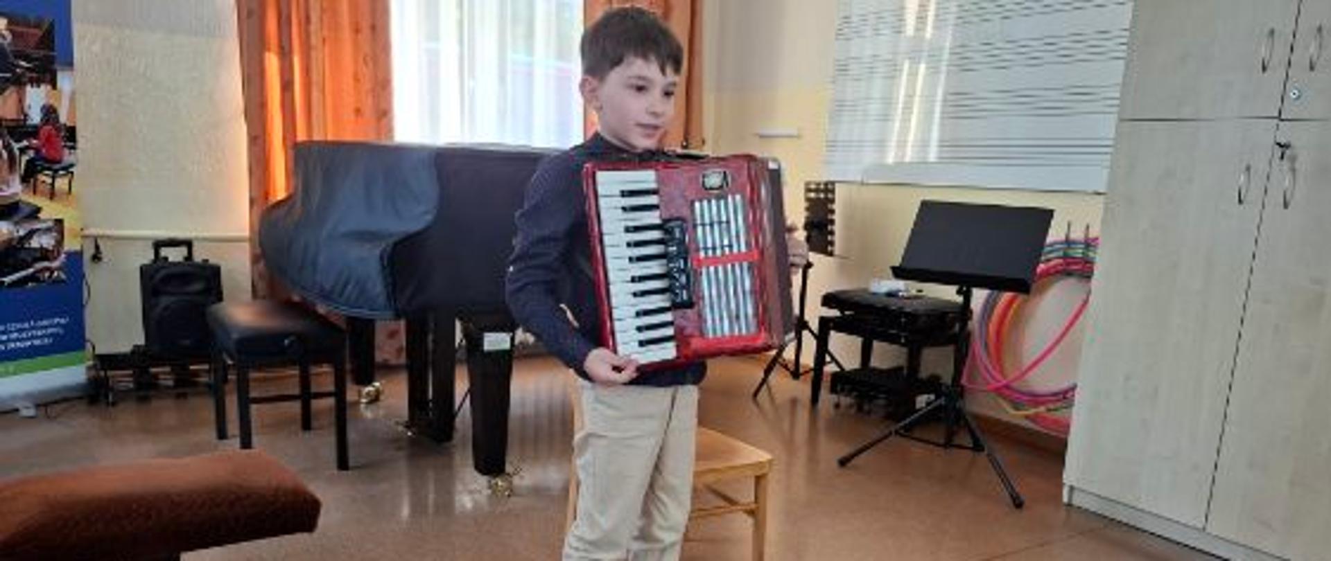 Na zdjęciu znajduje się młody chłopiec stojący w sali muzycznej i trzymający w rękach czerwony akordeon klawiszowy. Chłopiec ubrany jest w ciemną koszulę z długim rękawem, jasne spodnie oraz czarne buty sportowe. W tle widoczne jest wnętrze sali, w którym znajdują się instrumenty i akcesoria muzyczne: Duży czarny fortepian z nałożonym pokrowcem,Stołki muzyczne (w tym jeden tapicerowany na pierwszym planie), Stojak na nuty (pulpit), Szafka, hula-hop oparte o ścianę oraz tablica do zapisu nutowego.