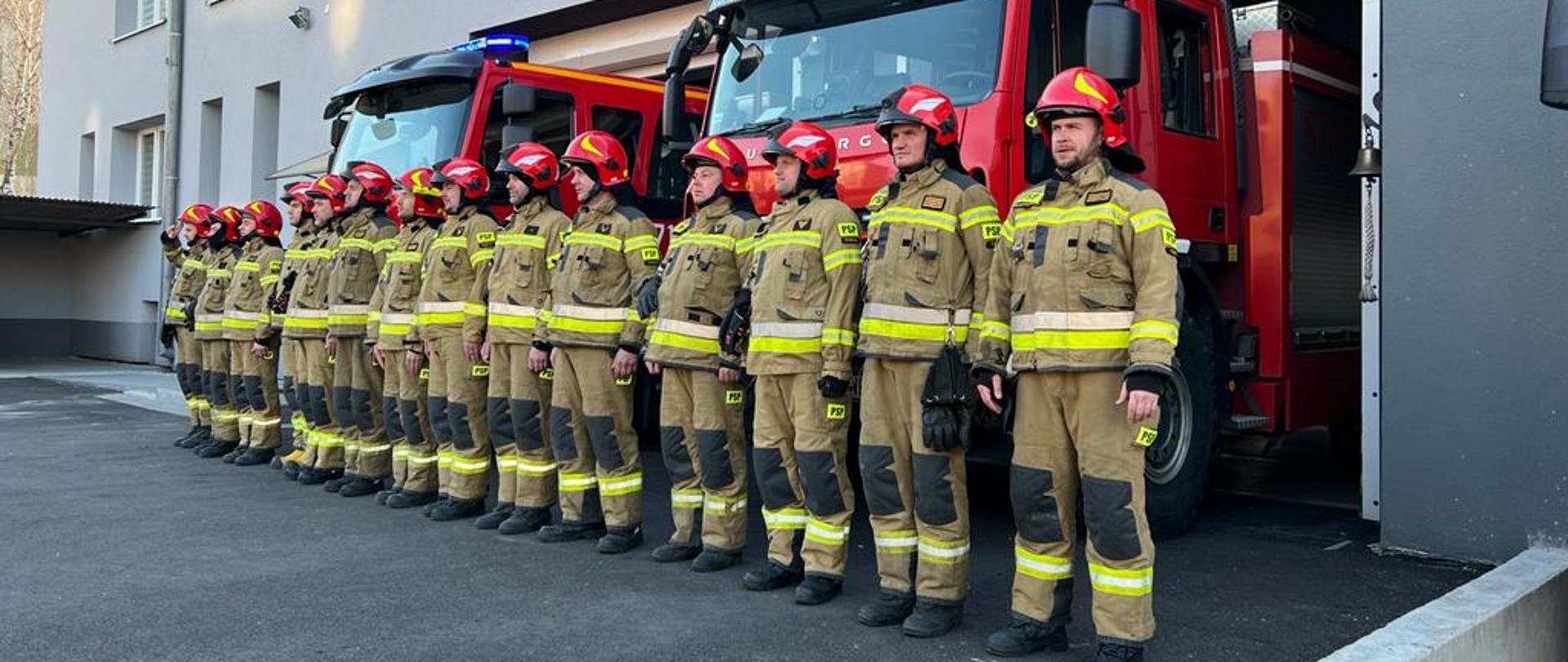 Strażacy ustawienie w rzędzie w mundurach bojowych na tle pojazdów