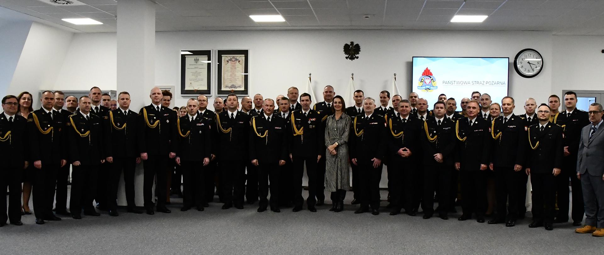 Strażacy pozują do zdjęcia grupowego wewnątrz pomieszczenia. 