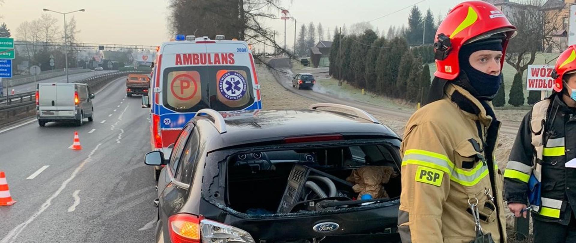 Na zdjęciu widoczny samochód osobowy z rozbitą tylną częścią oraz Zespół Ratownictwa Medycznego. Z prawej strony widoczni strażacy PSP w umundurowaniu specjalnym.