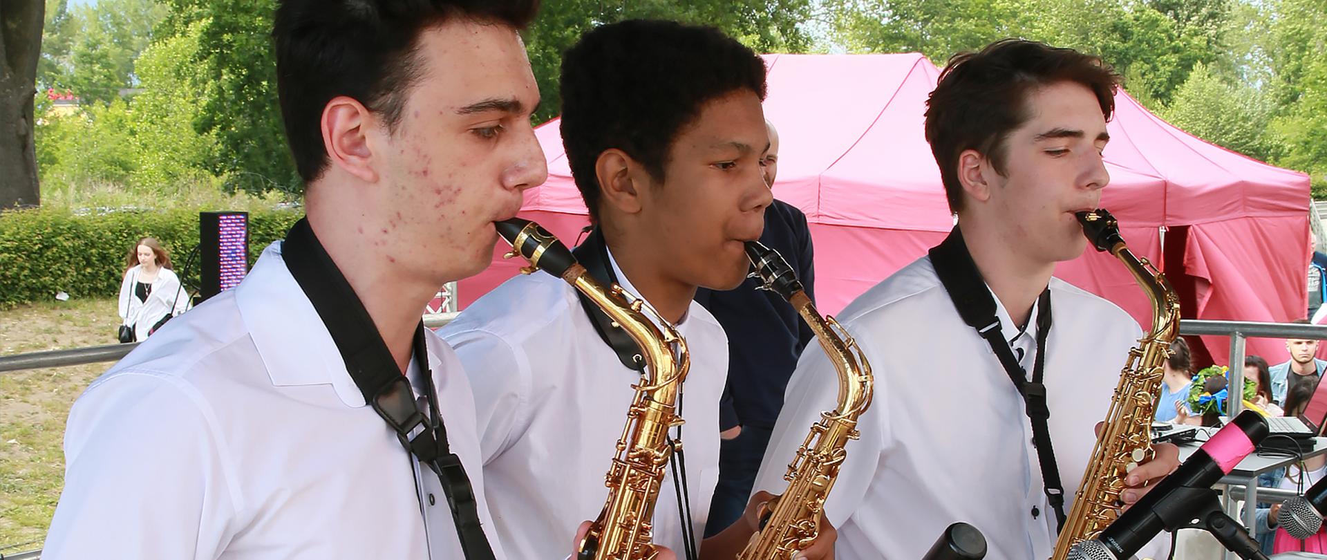 Na zdjęciu trzej chłopcy grają na saksofonach, w tle widoczny park i duży, czerwony namiot.