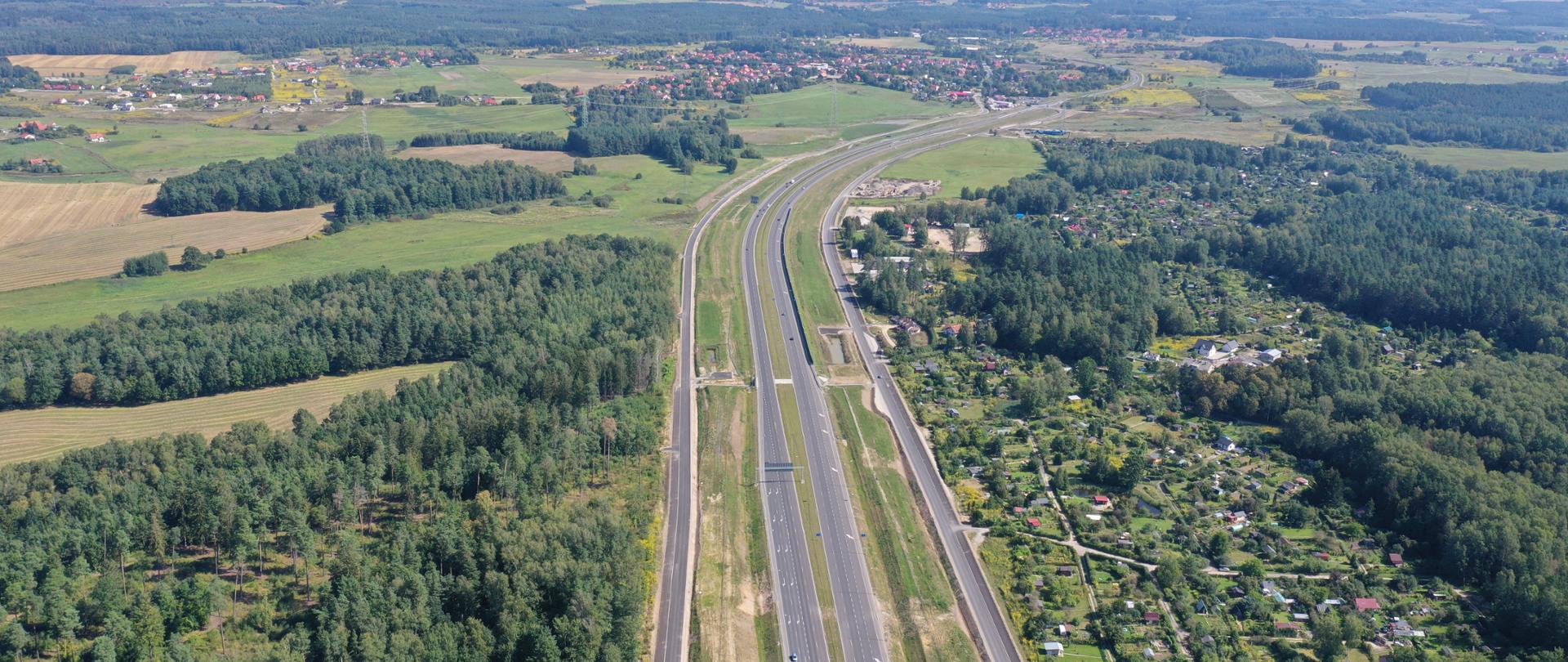 Dwujezdniowa droga ekspresowa. Po obu stronach znajdują się drogi serwisowe. Na horyzoncie zabudowania miejscowości.