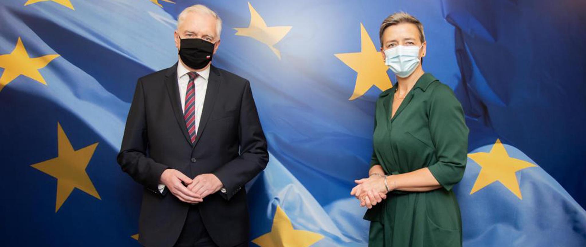 Wicepremier, minister rozwoju, pracy i technologii Jarosław Gowin w maseczce na twarzy, po jego prawej stronie stoi Wiceprzewodnicząca Wykonawcza Komisji Europejskiej ds. konkurencji Margrethe Vestager. Z tyłu flaga UE.