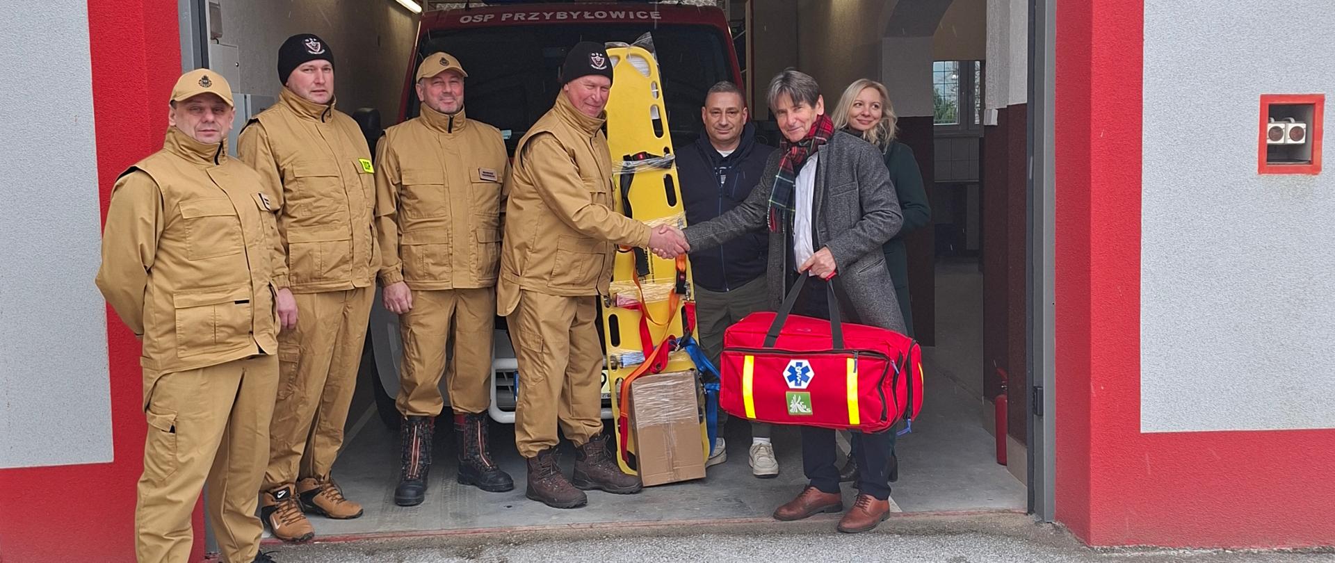 Grupowe zdjęcie siedmiu osób pozujących w bramie garażowej remizy Ochotniczej Straży Pożarnej w Przybyłowicach. Nad wejściem widnieje duży, czerwony napis "OSP PRZYBYŁOWICE". Czterech mężczyzn po lewej stronie ubranych w jasnobrązowe - beżowe mundury strażackie. W centrum kadru, mężczyzna w szarym płaszczu (Dyrektor Jan Socha) wręcza Prezesowi OSP Zdzisławowi Dąbrowskiemu (w mundurze) czerwoną torbę medyczną PSP R-1 i żółtą deskę ratowniczą. Obok nich, po prawej stronie, stoją: Kierownik Placówki Terenowej KRUS Sylwia Kaczorowska (w zielonym płaszczu), Pełnomocnik Marszałka Radosław Zysnarski. Wnętrze remizy, widoczne w tle, jest oświetlone i znajdują się tam fragmenty czerwonego wozu strażackiego.