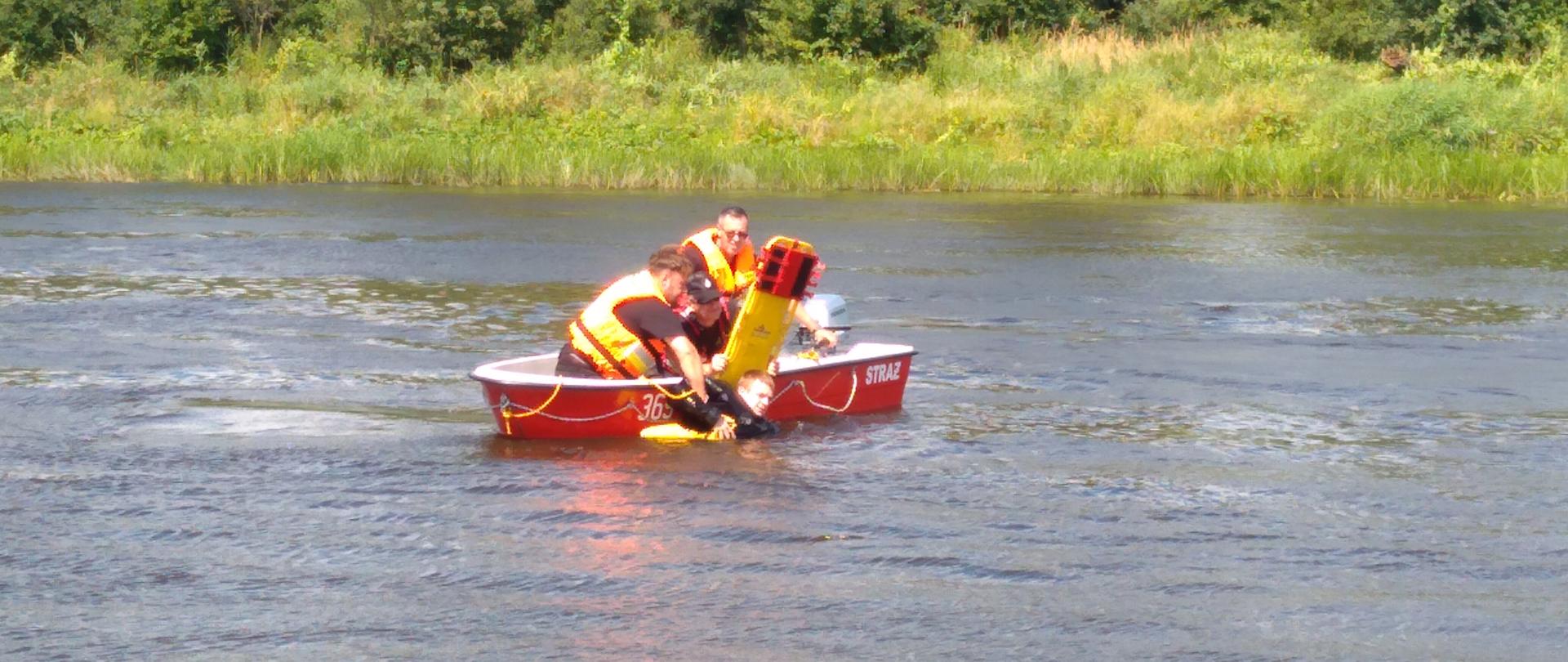 Trzech strażaków ubranych w kamizelki ratownicze którzy znajdują się na łodzi i podejmują osobę topiącą się przy pomocy deski ortopedycznej.