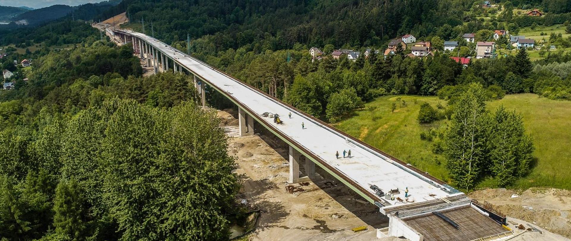 Budowa drogi ekspresowej S1 Przybędza - Milówka. Na zdjęciu widoczni pracownicy budowlani pracujący przy budowie estakady. Budowa przechodzi przez górzysty teren porośnięty gęsto zielenią. Estakada wznosi się ponad koronami drzew. W tle zabudowania mieszkaniowe.