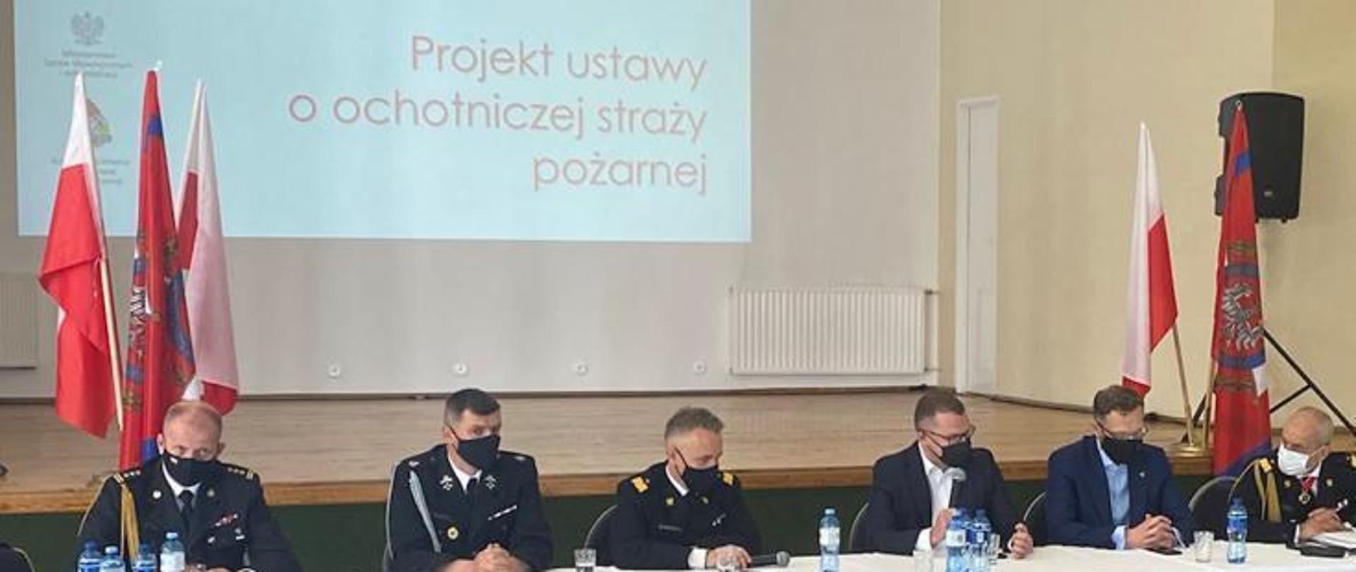 Przy stole siedzą mężczyźni - uczestnicy konsultacji, od lewej trzech strażaków, dwie osoby cywilne i kolejny strażak, w tle po lewo i prawo flagi a na wprost napis Projekt ustawy o PSP