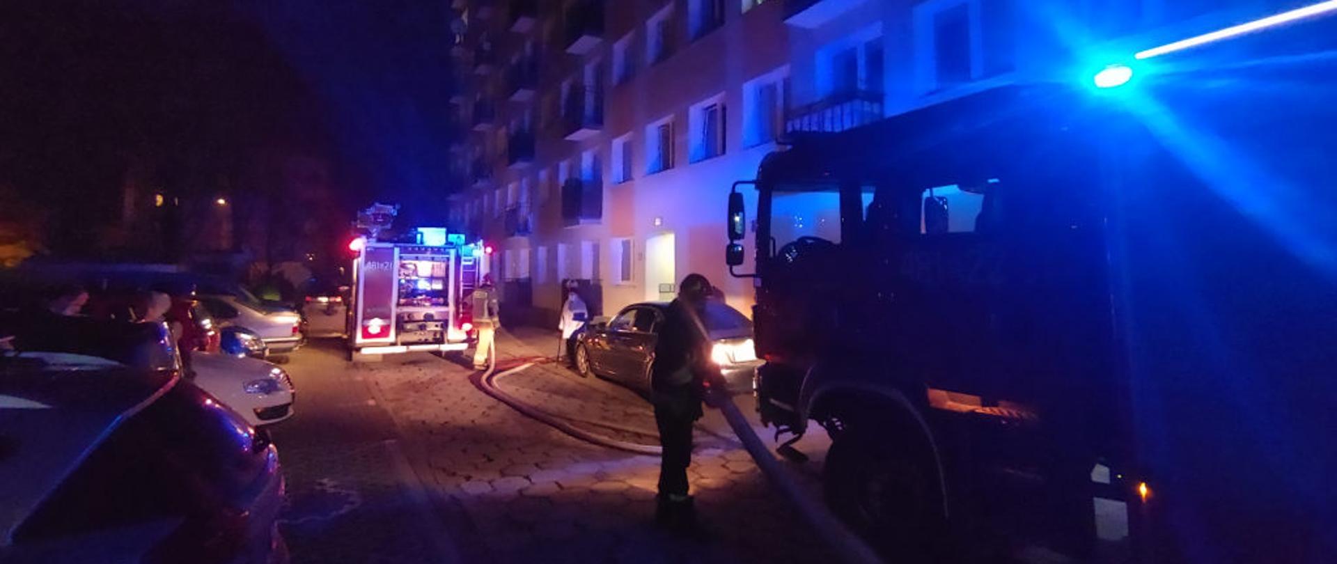 Zdjęcie zrobione w porze nocnej, w tle widać blok mieszkalny. Na pierwszym planie dwa pojazdy straży pożarnej, między nimi nawodnione linie gaśnicze i główne. Pomiędzy pojazdami dwóch strażaków. 