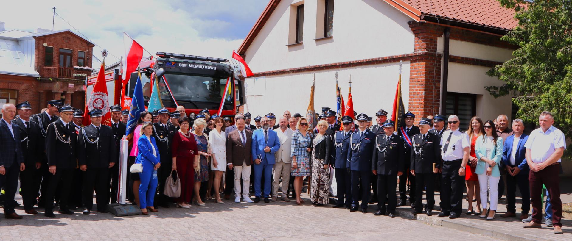 Zdjęcie przedstawia żywą, słoneczną scenę plenerową z czerwonym wozem strażackim, wyraźnie udekorowanym na ceremonię. Wóz jest ustawiony centralnie, przodem do widza. Jego przód zdobi duża czerwono-biała wstęga, rozciągnięta poziomo na całą szerokość, sugerująca ceremonię przecięcia wstęgi. Mniejsze czerwono-białe wstążki są zawieszone pionowo na masce, a dwie polskie flagi są umieszczone na masztach w górnych rogach kabiny, powiewające na wietrze.
Na górze kabiny wozu wyraźnie widoczne są napisy „OSP SIEMIĄTKOWO”. Pod przednim zderzakiem widoczna jest również tablica rejestracyjna „ZUL 6N7”. Sam wóz strażacki jest nowoczesny, z czerwoną karoserią i białymi akcentami na dolnej części przodu.