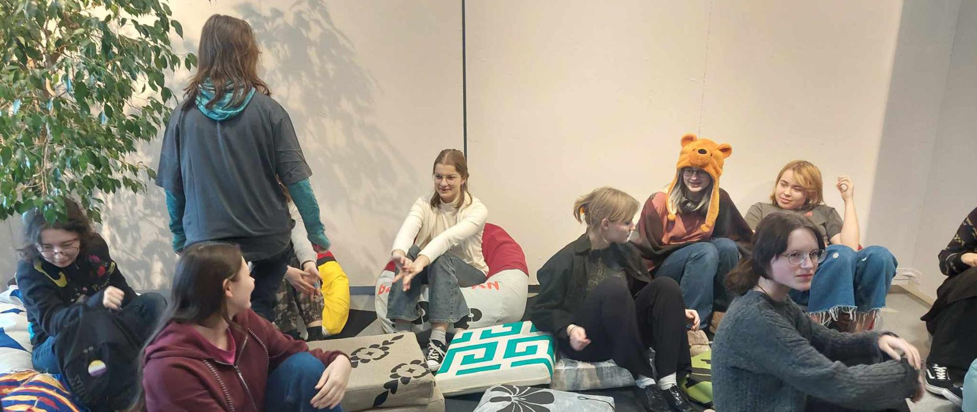 Fotografia. Białe ściany, grafitowe i szare wykładziny na podłodze. Kilka uczennic siedzi na poduszkach znajdujących się na podłodze w holu szkoły. Po lewej stronie stoi duży, doniczkowy kwiat.