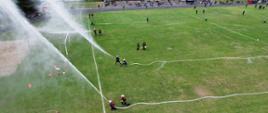 zdjęcie z lotu ptaka przedstawia stadion sportowy na murawie strażacy oraz rozwinięte dwie linie gaśnicze woda z prądownic tryska do góry