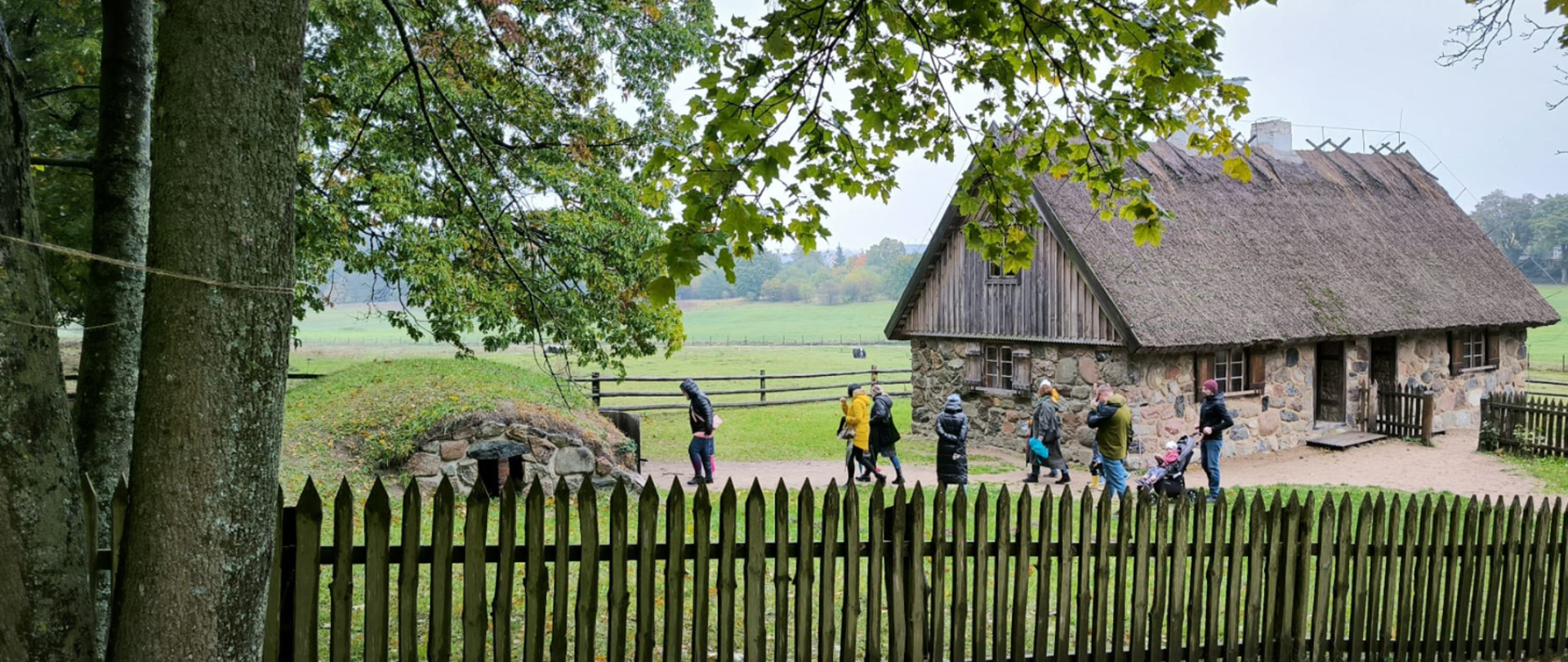 Na pierwszym planie drewniany płot, a w lewym rogu drzewo. Za ogrodzeniem grupa zwiedzających. Obok stary budynek, piwnica. W oddali łąki, las.