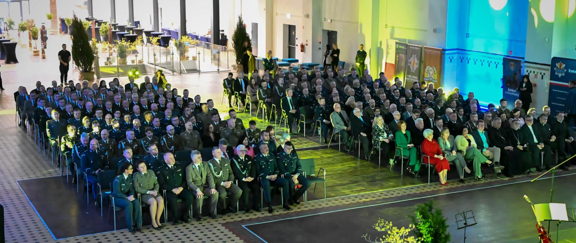 Szerokie ujęcie z góry na dużą salę wypełnioną rzędami krzeseł. Dominują osoby w mundurach. Po prawej stronie widać oświetloną scenę i fragment dekoracji.