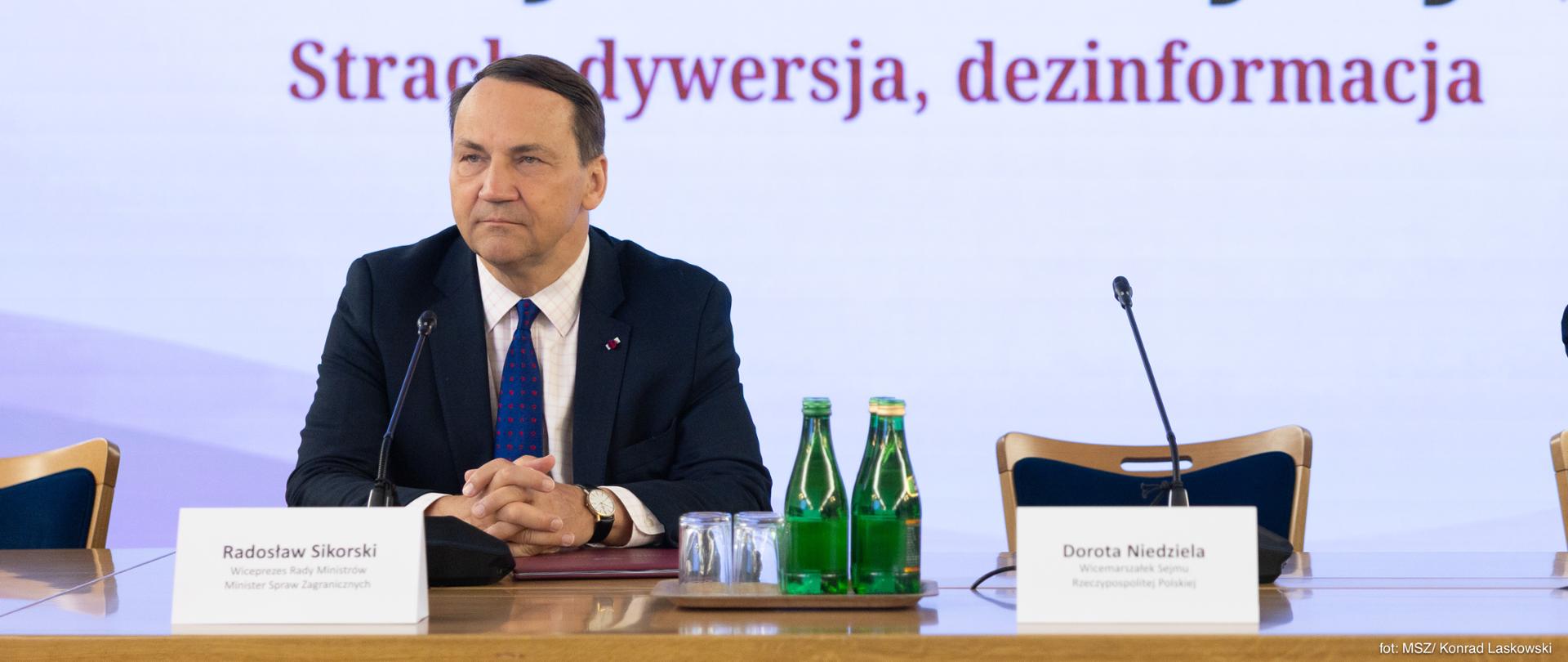 Wicepremier, minister spraw zagranicznych Radosław Sikorski siedzący przy stole prezydialnym przed ekranem z tytułem wydarzenia poświęconego zagrożeniom informacyjnym.