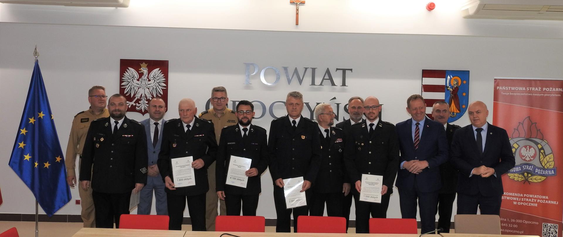 Strażacy stoją na tle ściany. Stoją w dwóch rzędach. Strażacy stojący z przodu trzymają w ręku dokumenty. z lewej strony strażaków stoi flaga unijna, a z prawej baner reklamowy z logo PSP. Na ścianie za strażakami jest napis powiat opoczyński a po obu jego stronach wisi Godło Polski oraz herb powiatu. Przed mężczyznami widzimy jasne stoły na których stoją mikrofony. Przed stołami stoją krzesła. 