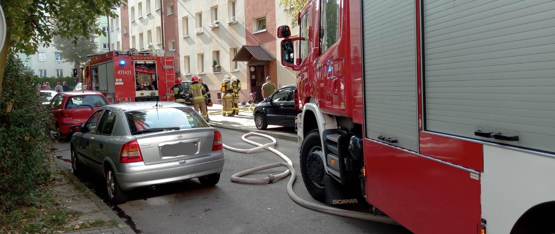 Zdjęcie przedstawia samochody strażackie i wykonane rozwinięcia linii gaśniczych podczas pożaru na osiedlu 1000-lecia w Myślenicach. W okolicy wejścia do klatki schodowej stoją strażacy. Na zdjęciu znajdują się również samochody osobowe zaparkowane na terenie osiedla.