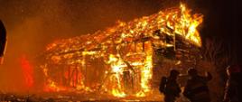 Na zdjęciu widoczna stodoła objęta w całości pożarem oraz trzech strażaków szykujących się do działań gaśniczych