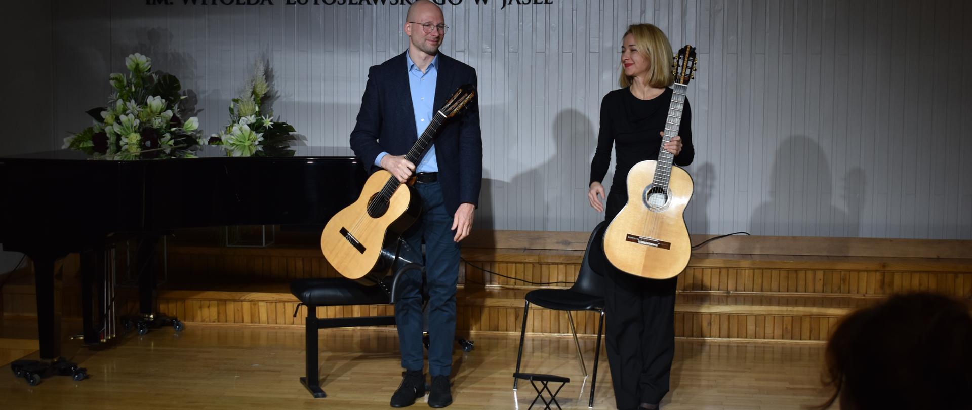 Pośrodku stoją ubrani w ciemne ubrania mężczyzna i kobieta oboje uśmiechnięci trzymają w rękach gitary. Obok nich stoją czarne krzesła i podnóżek gitarowy. Za nimi fragment czarnego fortepianu za którym widać biało-zielone kwiaty. W tyle jasna ściana na której widnieje logo szkoły dwie kolorowe nutki pośrodku z klawiszami fortepianu i napis Państwowa Szkoła Muzyczna I stopnia im. Witolda Lutosławskiego w Jaśle. Na ścianie widać również fragment godła państwowego.