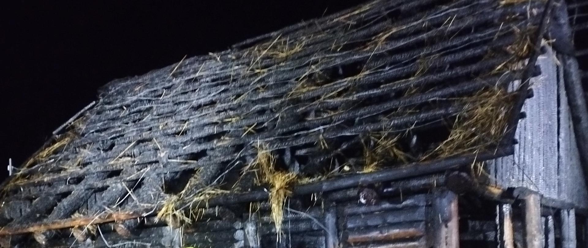Zdjęcie w porze nocnej. Widać nadpalony budynek drewniany. Podłoże trawiaste. Oświetlenie sztuczne.