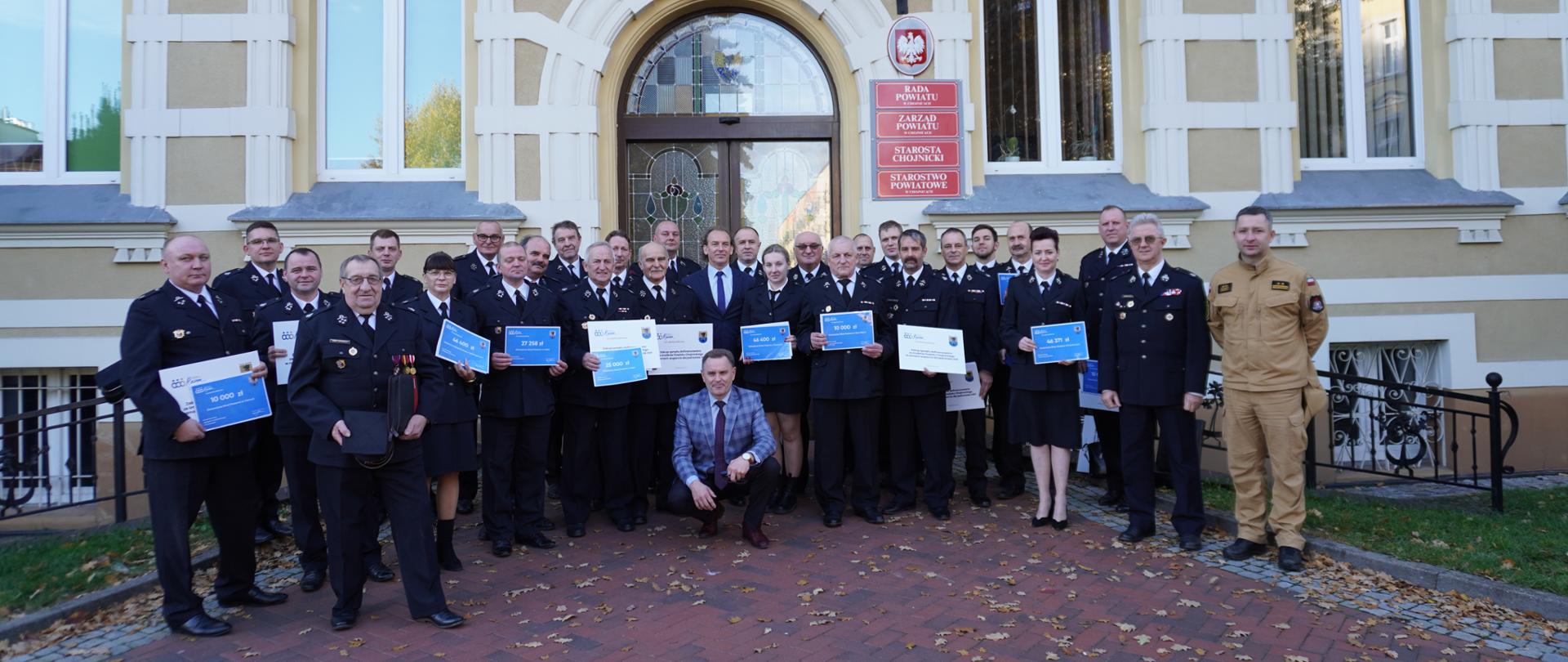 Zdjęcie grupowe przedstawiające grupę kilkudziesięciu osób. Większość z tych osób to strażacy druhowie OSP w umundurowaniu galowym. Zdjęcie wykonane przed wejściem głównym do budynku Starostwa powiatowego w Chojnicach. 