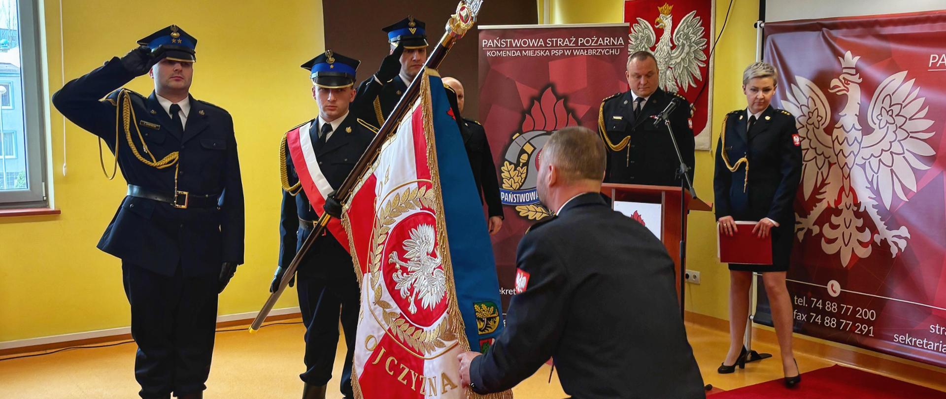 Strażacy mundurach wyjściowych stoją na baczność. Jeden ze strażaków trzyma w ręce niebieski sztandar. Komendant Miejski PSP w Wałbrzychu klęka na czerwonym dywanie przed sztandarem