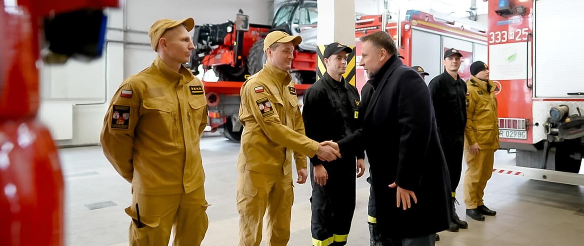 Mężczyzna w ciemnym płaszczu podaje rękę strażakowi w żółtym mundurze PSP. W tle stoją pozostali funkcjonariusze oraz wozy strażackie.