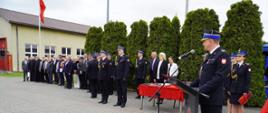 Komendant Powiatowy Państwowej Straży Pożarnej w Rypinie stoi przy mównicy i przemawia do uczestników uroczystości odbywającej się na placu przed jednostką straży pożarnej. W tle widoczni są strażacy w galowych mundurach, przedstawiciele władz, duchowieństwo oraz zaproszeni goście ustawieni wzdłuż placu. Obok przygotowano stół z odznaczeniami i dokumentami do wręczenia podczas ceremonii.