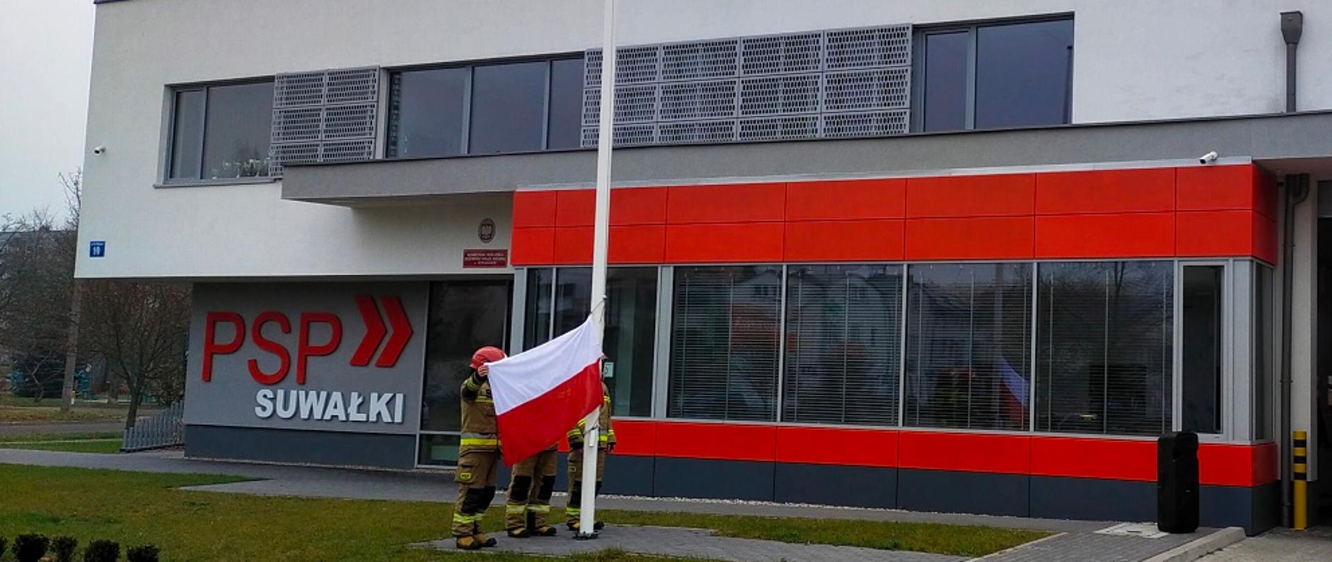 Zdjęcie obrazuje podniesie flagi państwowej na maszt przez strażaków w tle budynek KM PSP w Suwałkach