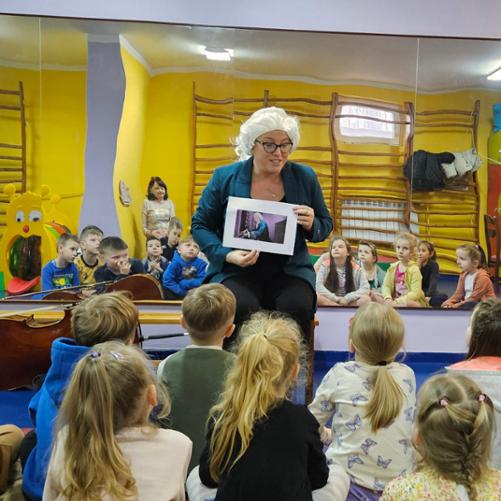 Zdjęcie przedstawia dzieci przysłuchujące się opowieści o Janie Sebastianie Bachu przedstawianej przez Panią Urszulę Właźniak. Pani Urszula trzyma w dłoniach grafikę przedstawiającą sylwetkę J. S.Bacha. Na głowie ma perukę barokową w kolorze białym. W tle lustra w których widać sylwetki dzieci.