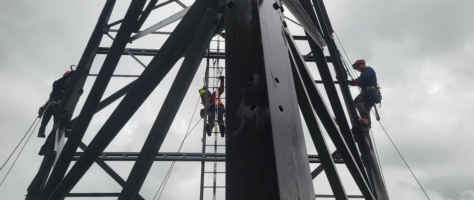 Ćwiczenia z zakresu ewakuacji z konstrukcji budowlanych, trzy osoby wykonują ćwiczenia znajdując się na maszcie kratownicowym