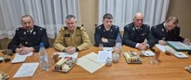Strażacy Państwowej Straży Pożarnej oraz ochotniczych straży pożarnych siedzą za stołem na którym są dokumenty oraz poczęstunek.