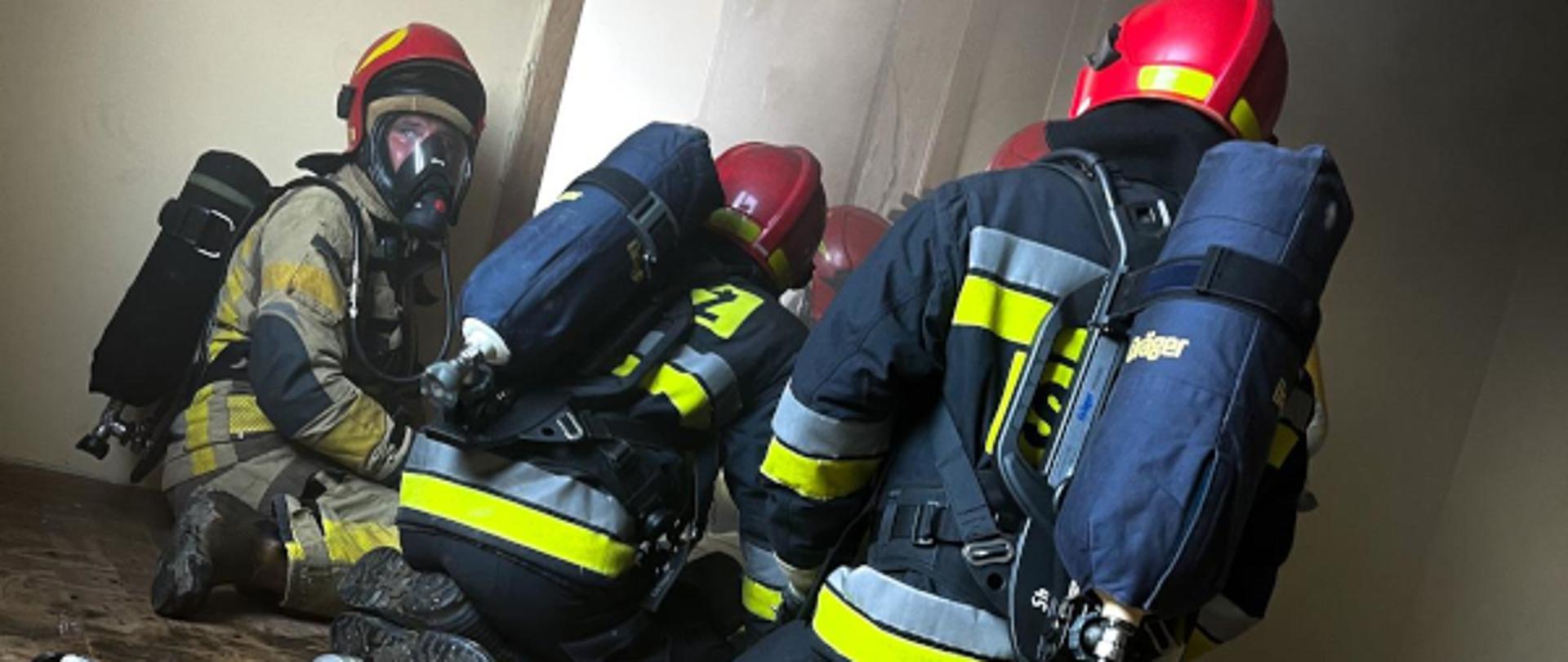 czterech strażaków ubranych w ubrania specjalne przygotowuje się do wejścia do pomieszczenia objętego pożarem, z drzwi wydobywa się dym