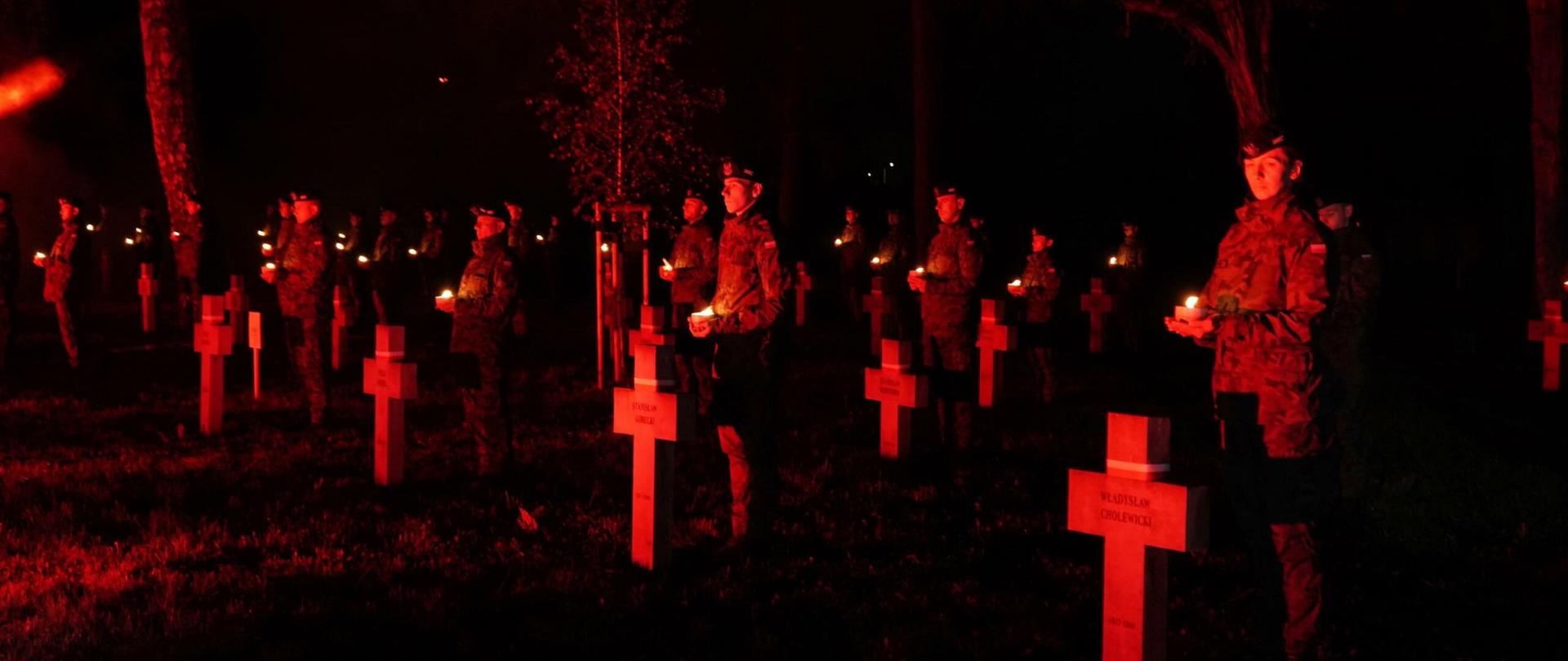 Żołnierze z zapalonymi zniczami stoją za krzyżami nagrobków.
