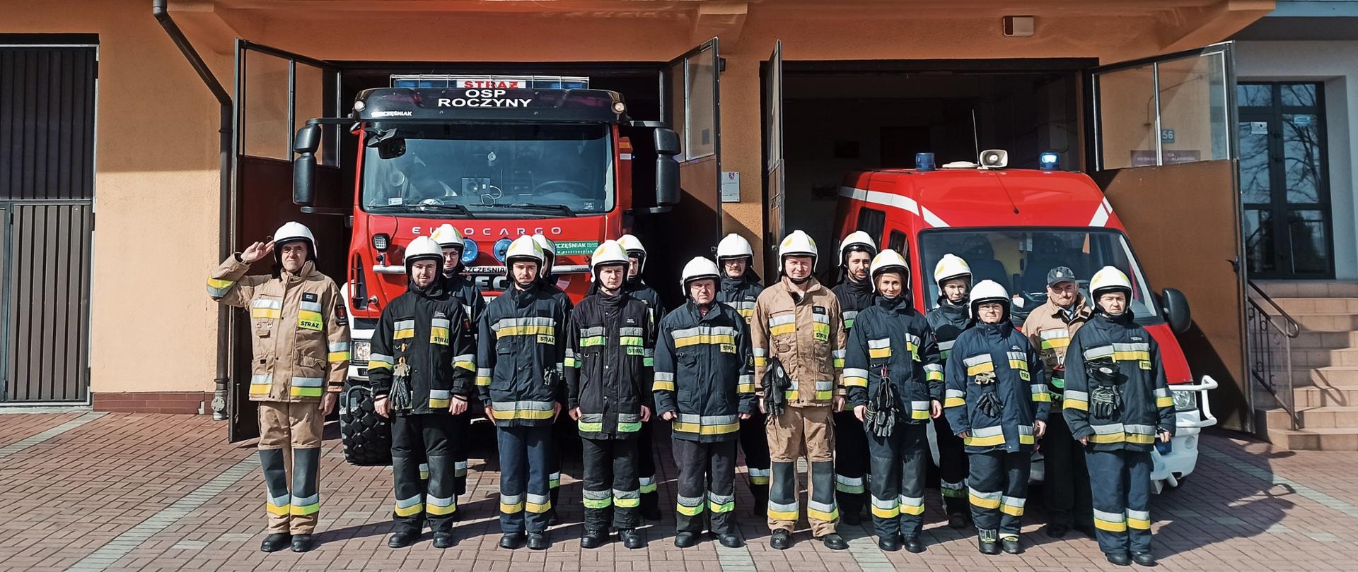 Widok z przodu. Szesnaścioro strażaków OSP Roczyny stoi w ubraniach specjalnych przed samochodami pożarniczymi przed garażem OSP. 
