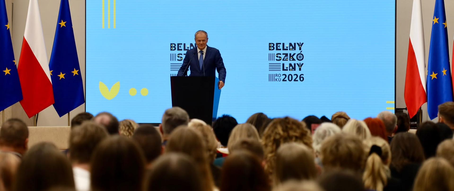 Na zdjęciu przedstawiono wystąpienie podczas oficjalnej konferencji. W centrum sceny stoi mężczyzna w garniturze, który przemawia przy ciemnej mownicy do licznie zgromadzonej publiczności.