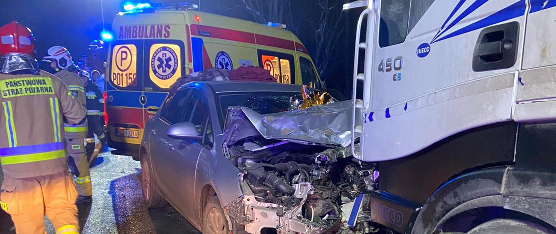 Srebrny samochód z uszkodzonym przodem, za nim karetka pogotowia